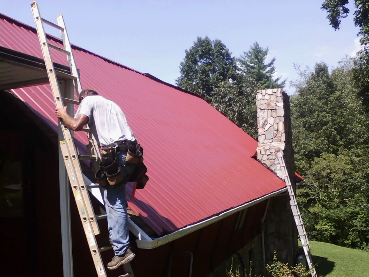 Roofer on a ladder beside a red metal roof and stone chimney — Soffit & Fascia Repair by Roofing Brothers in Corning
